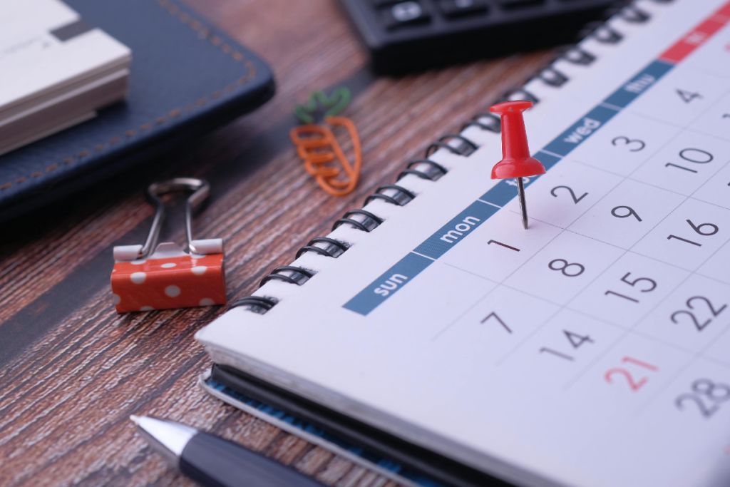A red pushpin marks Monday the 1st on a desk calendar surrounded by office supplies