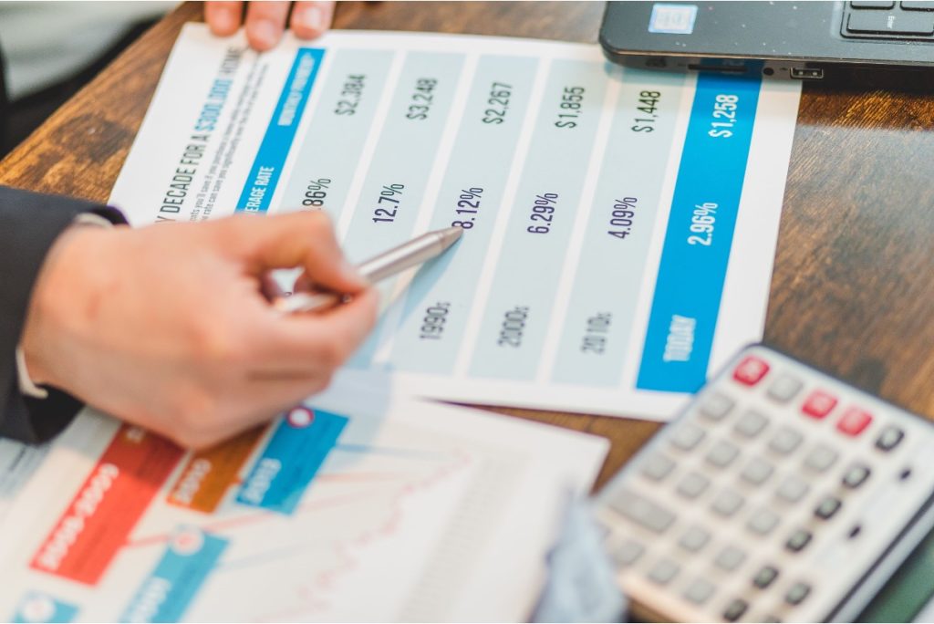A hand holds a pen over a rate comparison chart beside a calculator and laptop.
