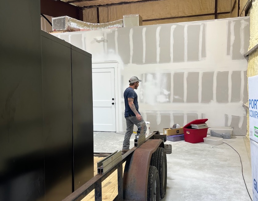 A man standing beside a trailer bed facing a freshly mudded drywall wall in a warehouse space