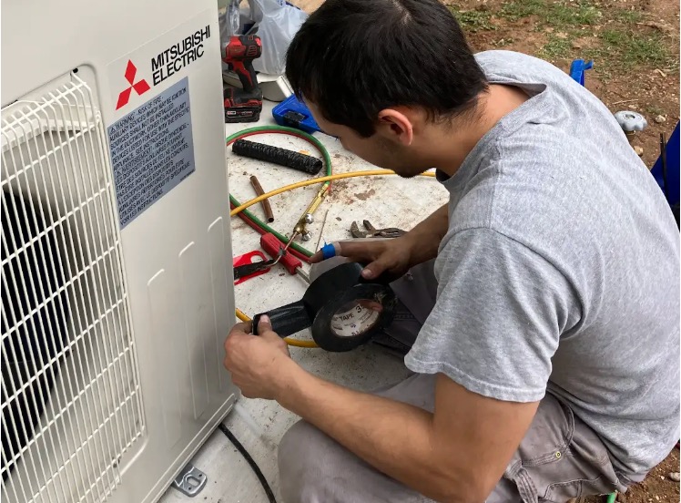 A technician uses black electrical tape while installing a Mitsubishi Electric outdoor air conditioning unit.