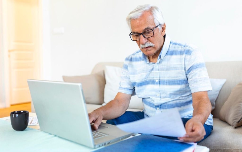 An older man in a striped shirt reviews paperwork while working on a laptop at home