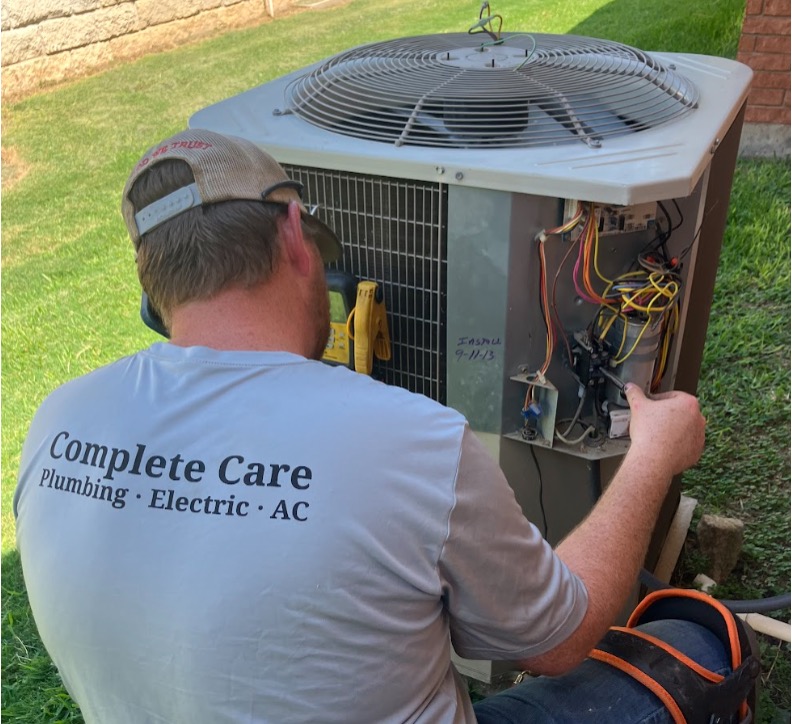 HVAC technician from Complete Care Plumbing Electric AC, servicing an outdoor air conditioning condenser unit, inspecting electrical components and wiring during a maintenance or repair visit. 