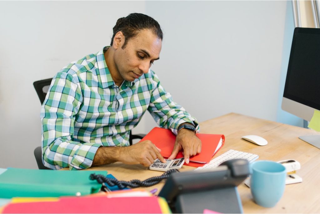 Man in a plaid shirt uses a calculator at a desk with a red folder and a computer.