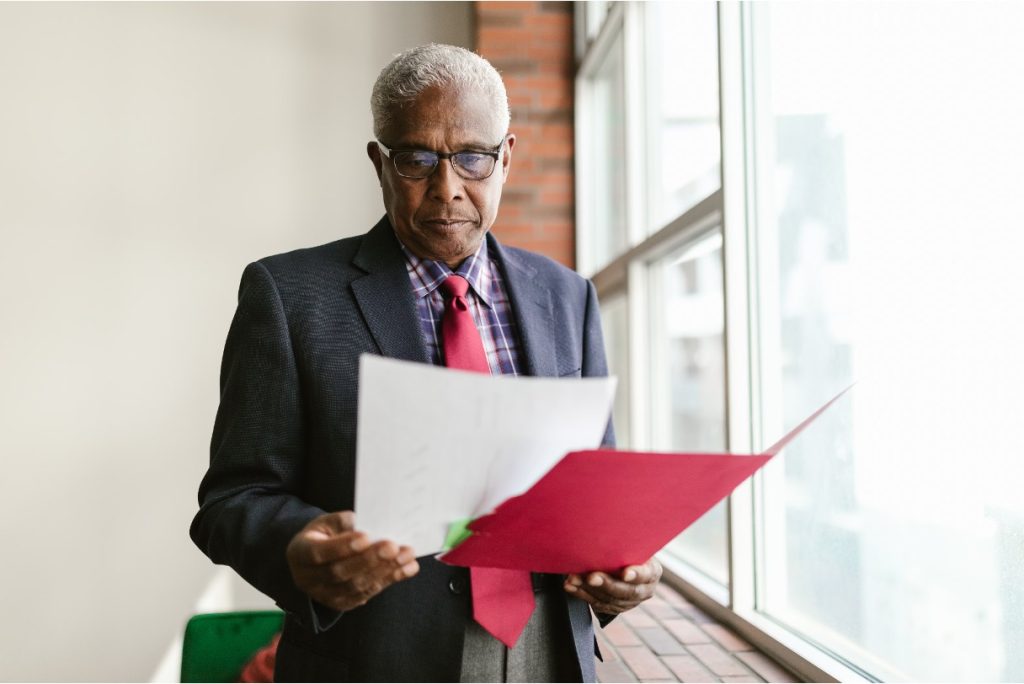 Older man in a suit and red tie reads documents from a red folder near a window.