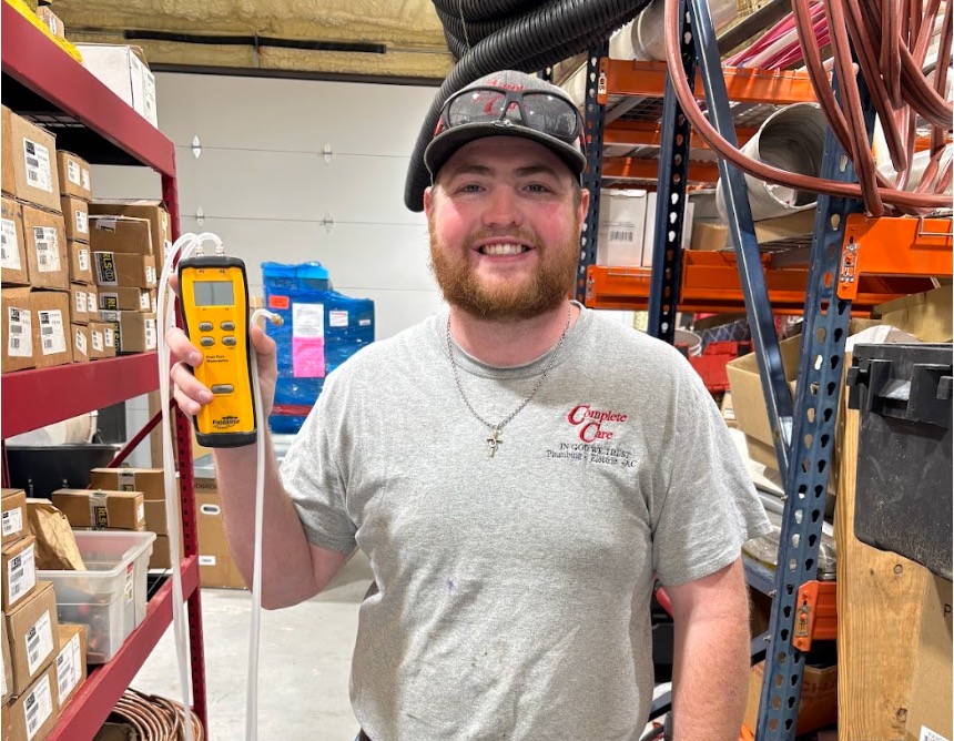Smiling bearded man in a warehouse holds a yellow handheld gas detector, wearing a "Complete Care" shirt