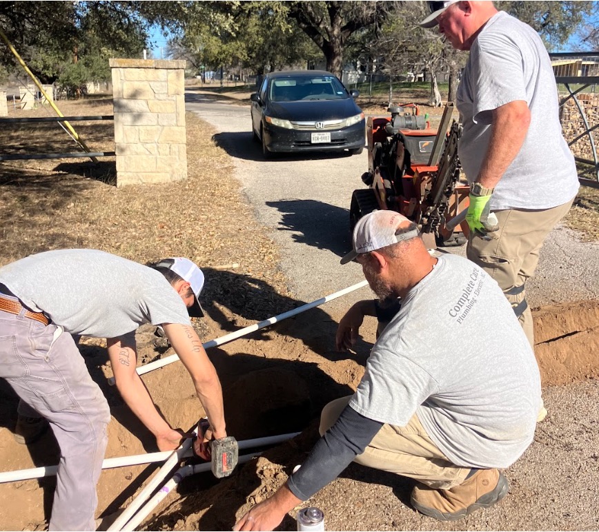 Three workers install PVC piping in a dug hole near a driveway and a parked car.
