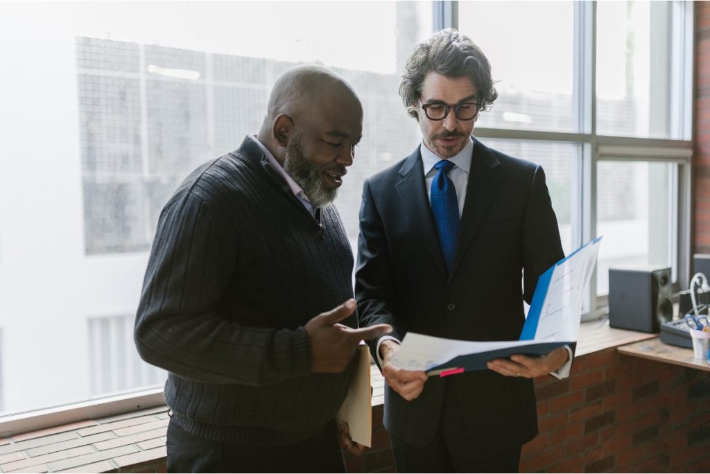 Two men review documents in a folder together while standing near a large office window.