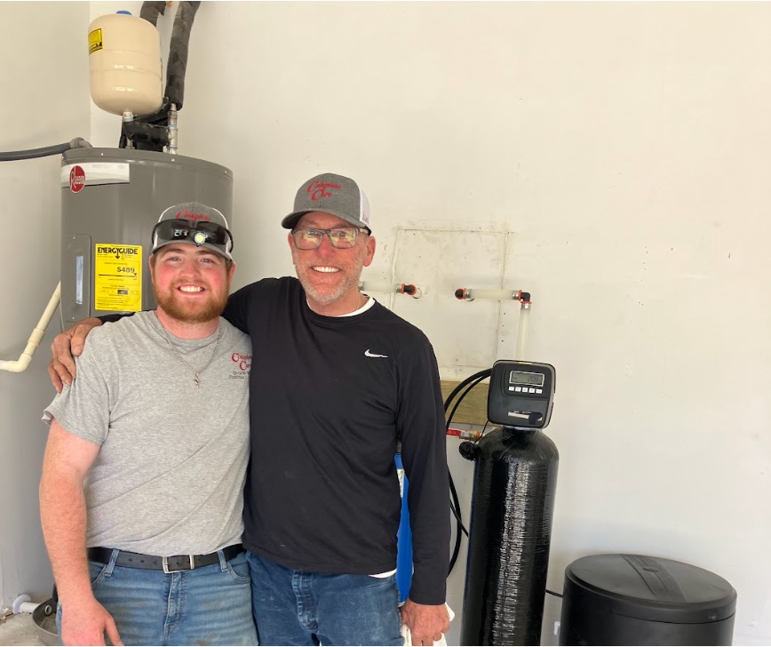 Two smiling men stand arm in arm beside a water heater and water softener system