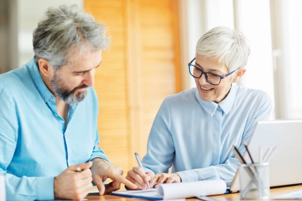 Two smiling older adults in blue shirts review documents together at a desk with a laptop.