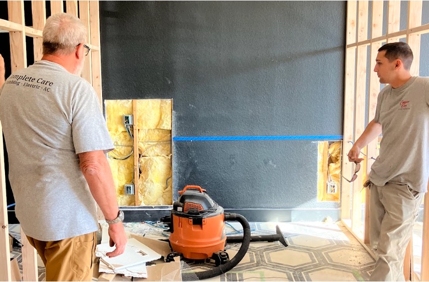 Two tradesmen in a room under renovation with exposed insulation, wall framing, and an orange shop vacuum.