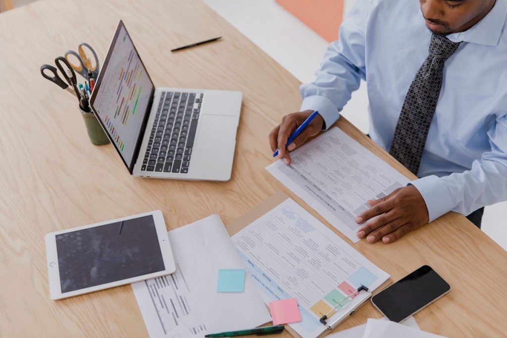 Man at a desk marking up printed schedules with a laptop and tablet beside him