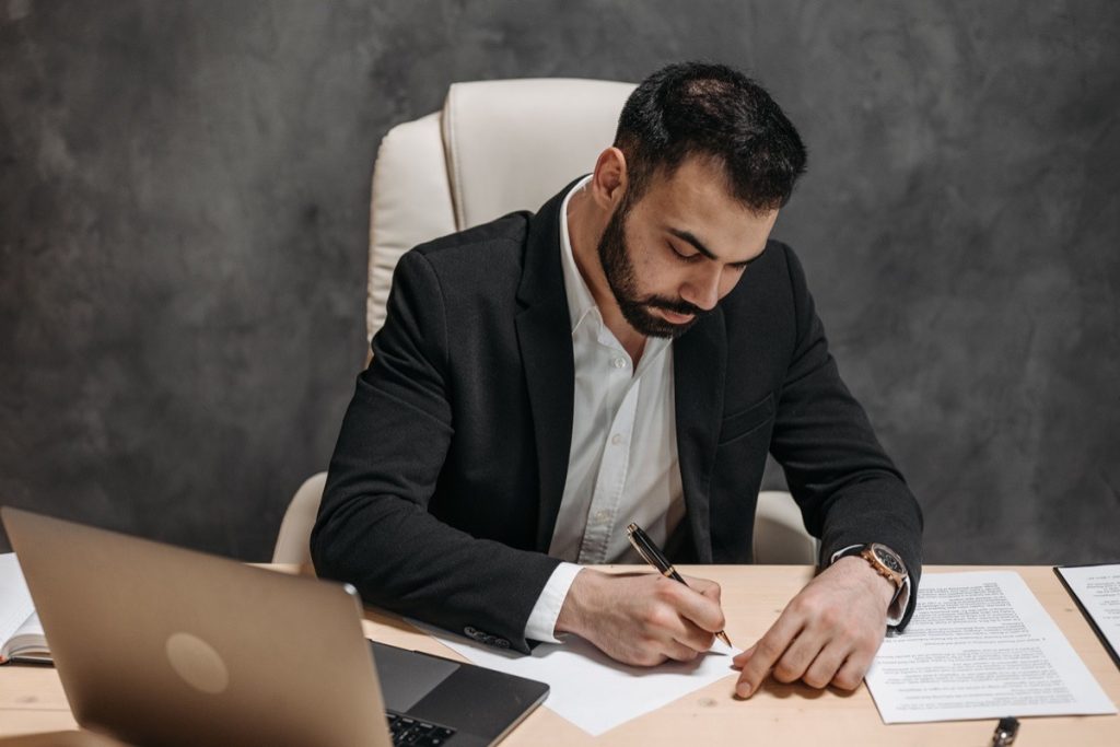 Man at a desk signing a document with contract pages spread beside him