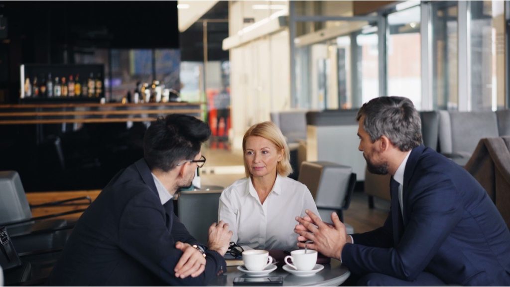 Three people in a serious discussion over coffee about earnout risks.