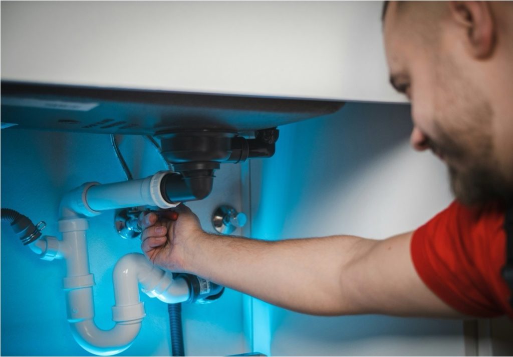 A plumber tightening pipe fittings under a sink during a residential service call.