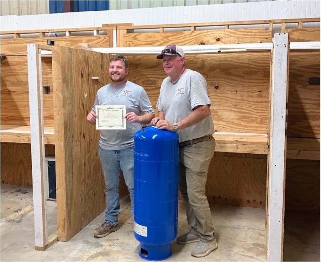 A plumbing professional holding a certificate while standing with a colleague beside a pressure tank in a workshop