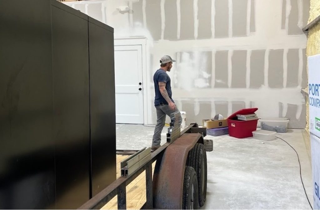 A tradesman standing in a warehouse workspace next to a trailer and supplies