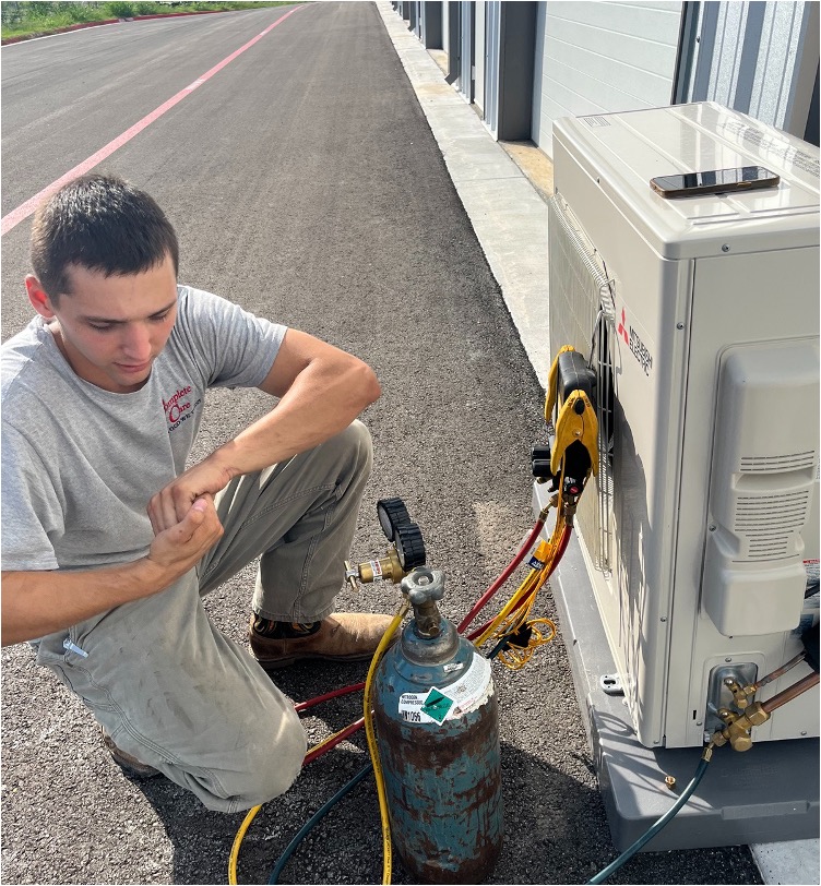 An HVAC technician beside a Mitsubishi condenser unit with a nitrogen tank and refrigerant lines connected
