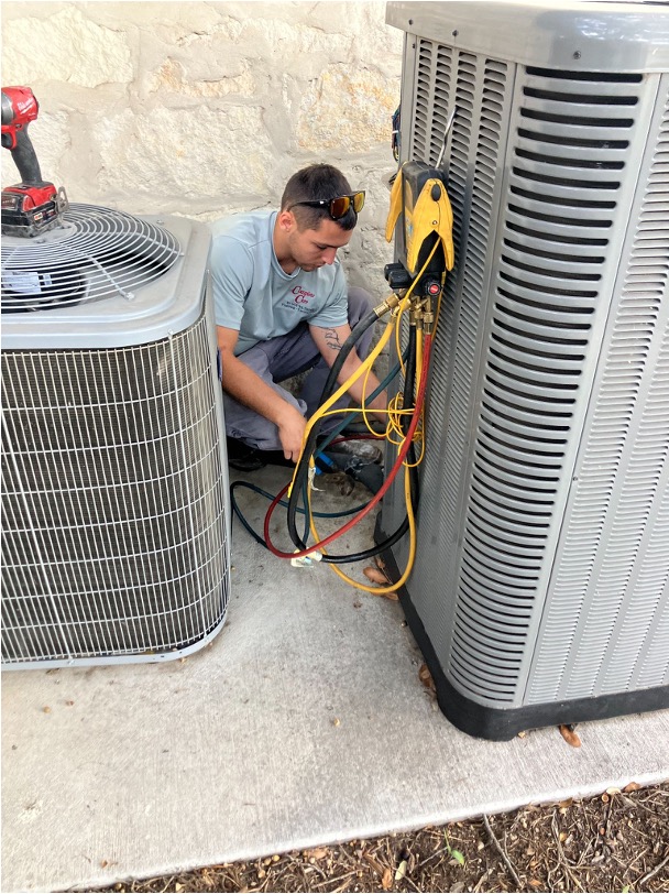 An HVAC technician working between two residential condenser units with refrigerant lines and tools