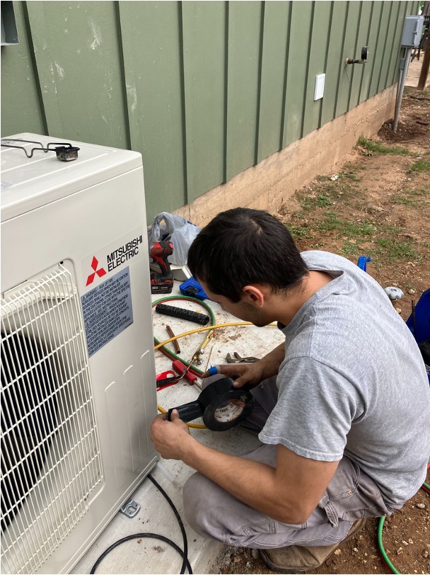 An HVAC technician working on a Mitsubishi Electric condenser unit with tools and tape