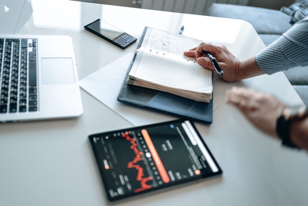 Hands working through figures in a notebook beside a laptop, calculator, and tablet