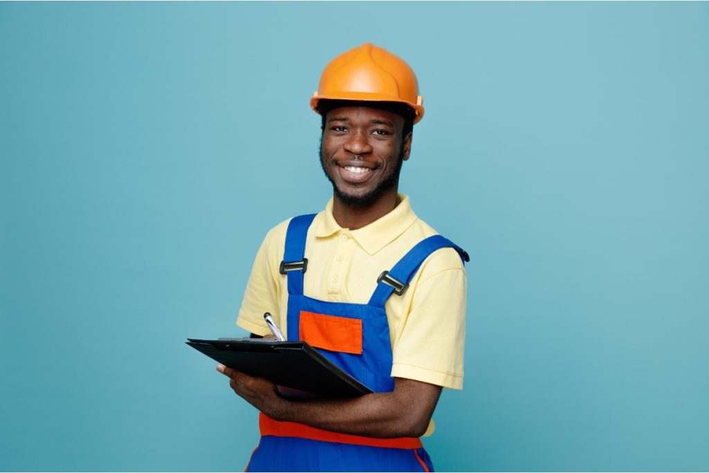 Smiling HVAC technician in orange hard hat and blue overalls holding a clipboard and pen. (Image courtesy of Magnific)
