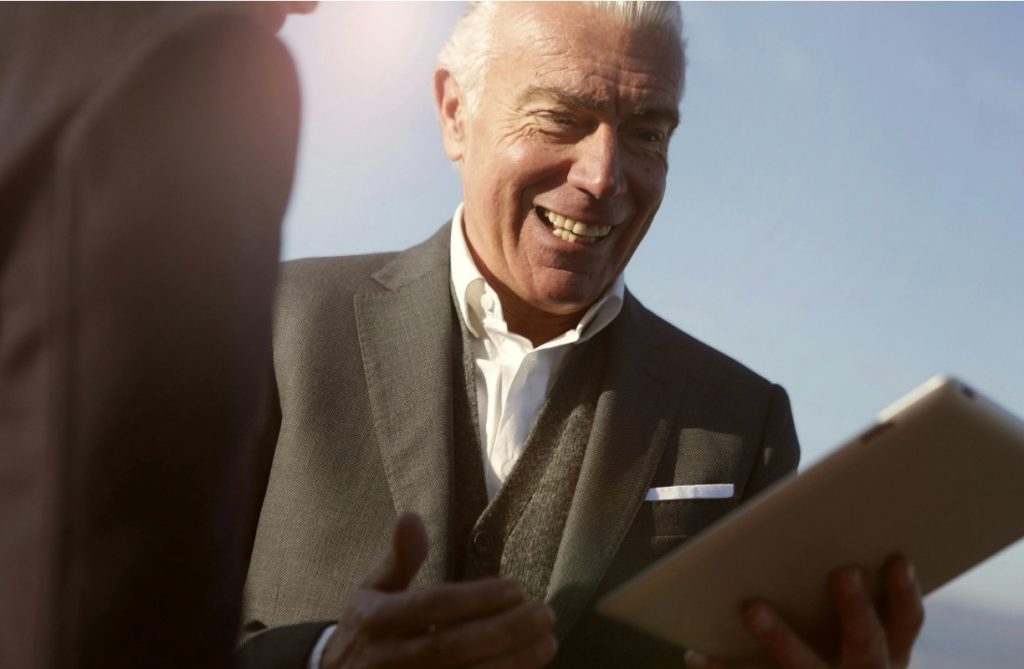 Smiling businessman in a suit holding a tablet while speaking with another person outdoors