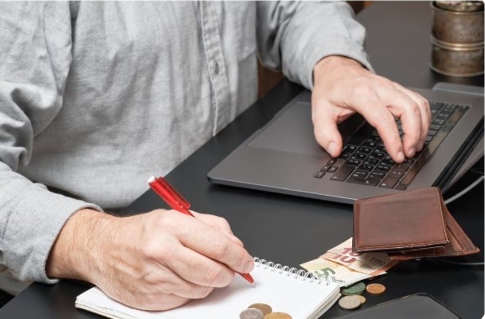 A person writing notes in a notebook while working on a laptop, with a wallet and cash on the desk