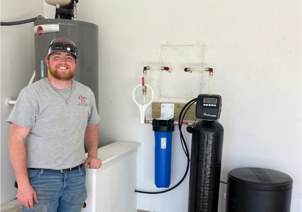 A smiling HVAC technician poses beside a completed water softener and filtration system installation indoors