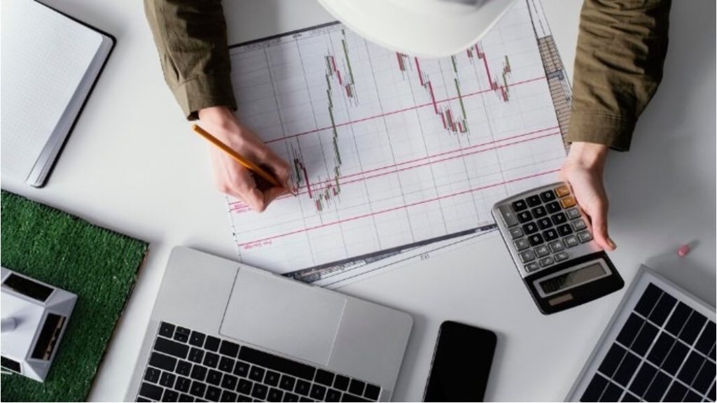 Overhead view of a person analyzing a stock market candlestick chart with a calculator and laptops nearby