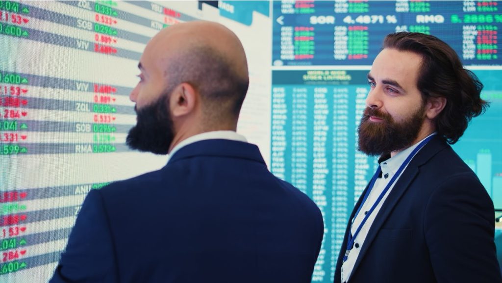 Two professionals standing before a live stock market ticker display.